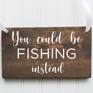 May include: A brown wooden sign with white text that reads "You could be FISHING instead". The sign is hanging from a white ribbon.