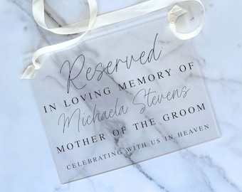 May include: A clear acrylic sign with the text "Reserved in loving memory of Michaela Stevens, Mother of the Groom, Celebrating with us in Heaven".