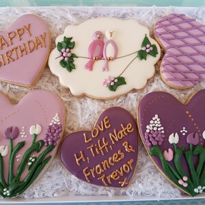 May include: A box of six heart-shaped cookies decorated with purple and gold icing. The cookies feature a variety of designs, including flowers, birds, and the words "Happy Birthday" and "Love H, Tiff, Nate Frances & Trevor".