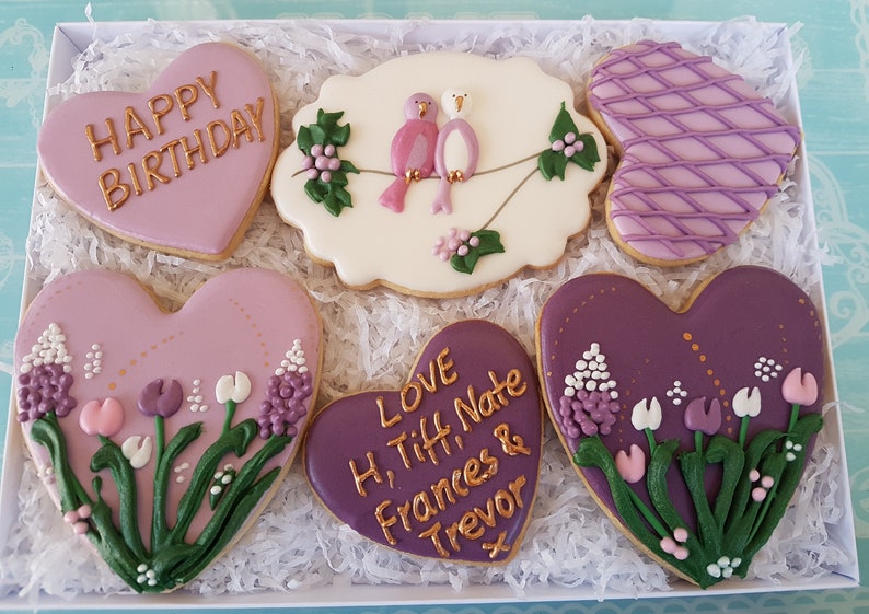 May include: A box of six heart-shaped cookies decorated with purple and gold icing. The cookies feature a variety of designs, including flowers, birds, and the words "Happy Birthday" and "Love H, Tiff, Nate Frances & Trevor".
