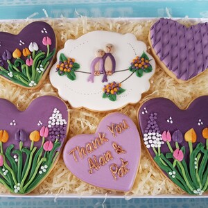 May include: A gift box filled with six heart-shaped cookies decorated with purple icing, flowers, and birds. The cookies are arranged in a box with shredded paper. One cookie has the text "Thank You Alan & Pat" in gold lettering.