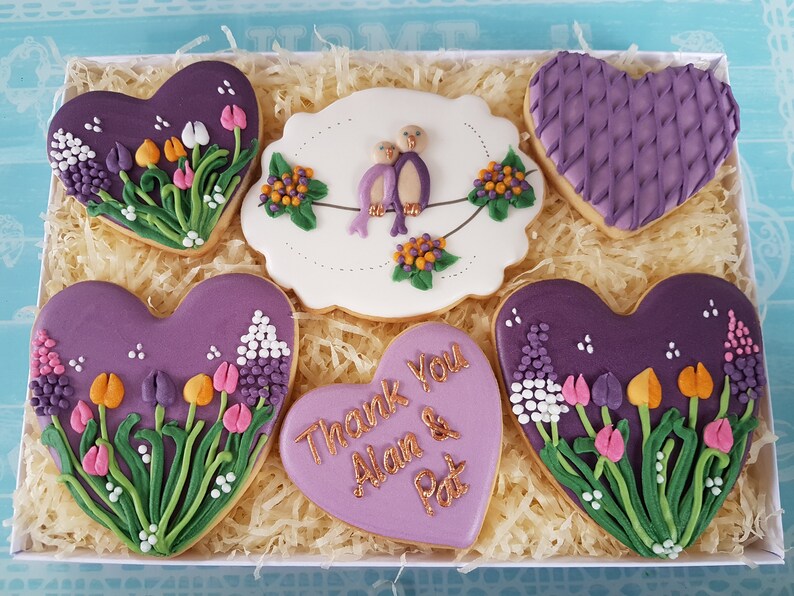 May include: A gift box filled with six heart-shaped cookies decorated with purple icing, flowers, and birds. The cookies are arranged in a box with shredded paper. One cookie has the text "Thank You Alan & Pat" in gold lettering.