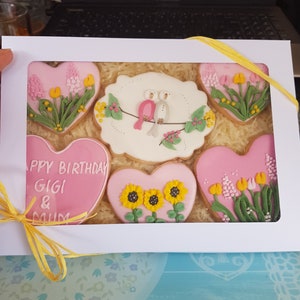May include: A white gift box with a yellow ribbon contains six pink heart-shaped cookies decorated with flowers and birds. The cookies say "Happy Birthday Gigi & Mum".