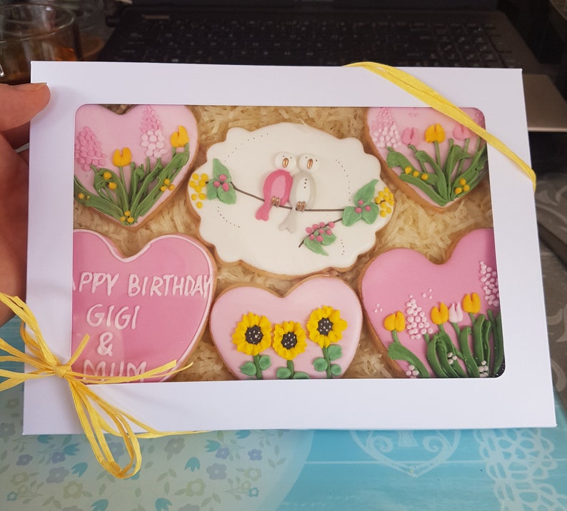 May include: A white gift box with a yellow ribbon contains six pink heart-shaped cookies decorated with flowers and birds. The cookies say "Happy Birthday Gigi & Mum".