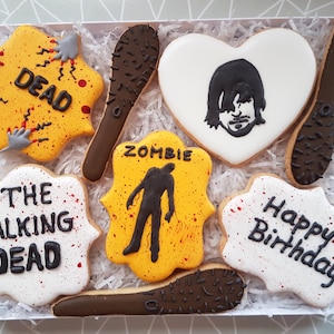 May include: A set of six decorated cookies in a white box with white shredded paper. The cookies are decorated with images and text related to the TV show "The Walking Dead". The cookies include a zombie, a barbed wire bat, a heart-shaped cookie with a character from the show, and a cookie that says "Happy Birthday".