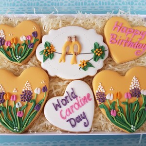 May include: A gift box filled with yellow heart-shaped cookies decorated with flowers and birds. The cookies have pink and purple icing and the text "Happy Birthday" and "World Caroline Day".