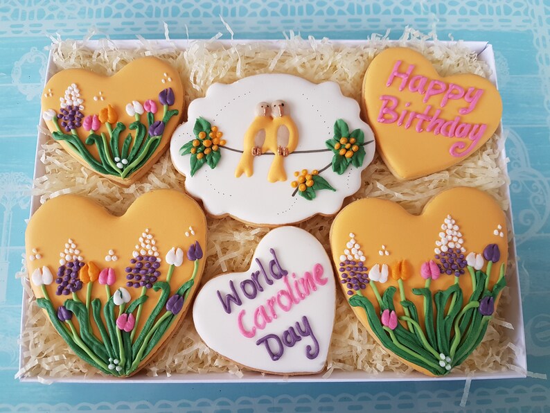 May include: A gift box filled with yellow heart-shaped cookies decorated with flowers and birds. The cookies have pink and purple icing and the text "Happy Birthday" and "World Caroline Day".