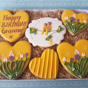 May include: A gift box filled with six heart-shaped cookies decorated with yellow icing, flowers, and birds. One cookie says "Happy Birthday Granny!" in gold lettering.