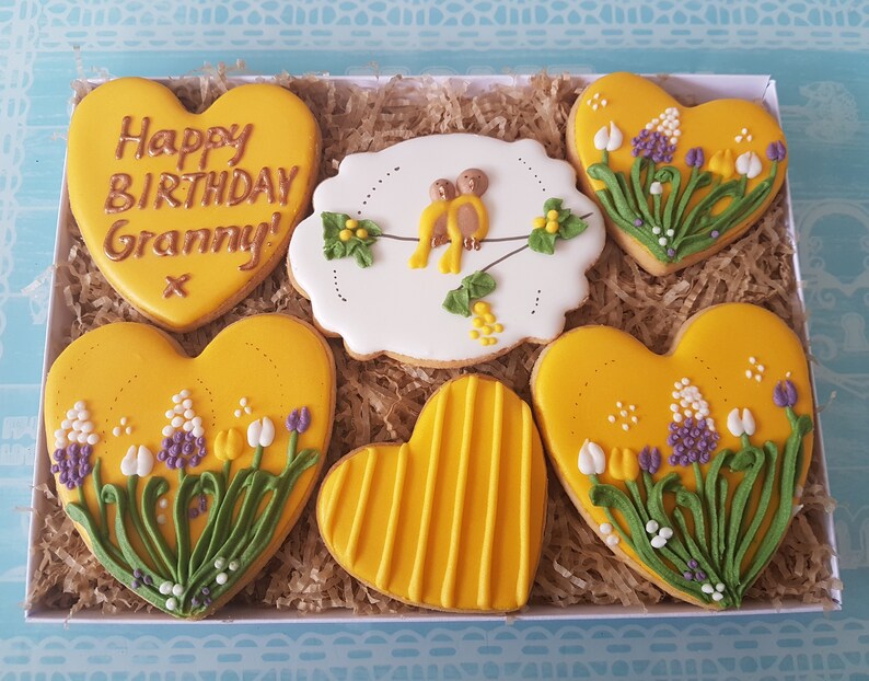 May include: A gift box filled with six heart-shaped cookies decorated with yellow icing, flowers, and birds. One cookie says "Happy Birthday Granny!" in gold lettering.