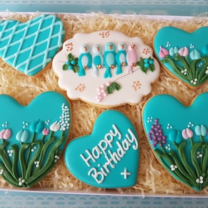 May include: A box of six heart-shaped cookies decorated with blue, pink, and white icing. The cookies feature a variety of designs, including birds, flowers, and the words "Happy Birthday".