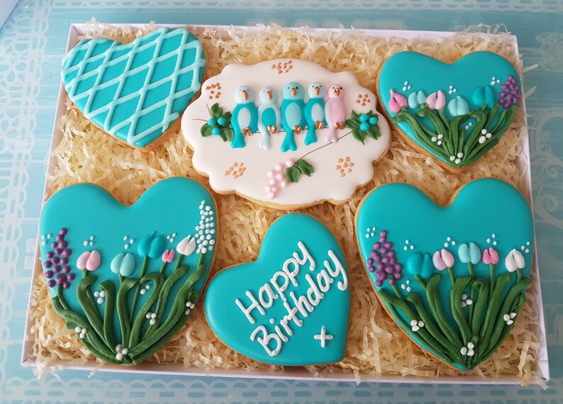 May include: A box of six heart-shaped cookies decorated with blue, pink, and white icing. The cookies feature a variety of designs, including birds, flowers, and the words "Happy Birthday".