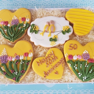 May include: A gift box filled with six heart-shaped cookies decorated with yellow icing and floral designs. The cookies feature a variety of floral patterns, including tulips, lilies, and sprigs of greenery. One cookie has the text "Happy 50th Anniversary" written in gold icing.