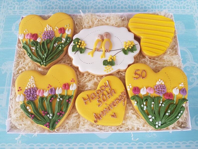 May include: A gift box filled with six heart-shaped cookies decorated with yellow icing and floral designs. The cookies feature a variety of floral patterns, including tulips, lilies, and sprigs of greenery. One cookie has the text "Happy 50th Anniversary" written in gold icing.