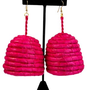 May include: A pair of vibrant fuchsia earrings. Each earring features a woven, beehive-shaped design suspended from a gold-colored hook with a twisted pink cord. The earrings are displayed on a black stand against a white background.