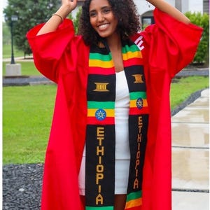 May include: A person wearing a red graduation gown and cap, holding the cap in their hand. They are wearing a white dress and a black and gold graduation stole with the word "ETHIOPIA" written on it.