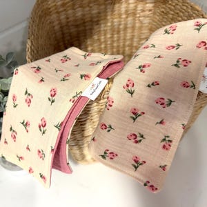Puede incluir: Dos paños de eructo rectangulares de tela beige claro con un patrón repetitivo de pequeños capullos de rosa y hojas verdes. Cada paño tiene un reverso de color rosa viejo sólido y una pequeña etiqueta blanca BabySweetCloth.