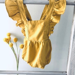 May include: Mustard yellow baby romper with ruffled sleeves and straps. The one-piece garment is made of a soft fabric and features a square neckline and a gathered waist. The romper is hanging on a gray ladder.