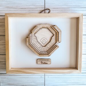 May include: A wooden model of Citizens Bank Park, home of the Philadelphia Phillies, in a light wood frame. The model is a top-down view of the baseball stadium.