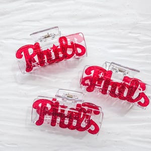 May include: Three clear plastic hair clips with the word "Phils" in red glitter script. The clips are rectangular with a hinged closure. The background is a textured white surface.