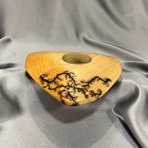 May include: A triangular wooden vase with a natural wood finish and a black lightning bolt design. The vase has a small, round opening at the top.