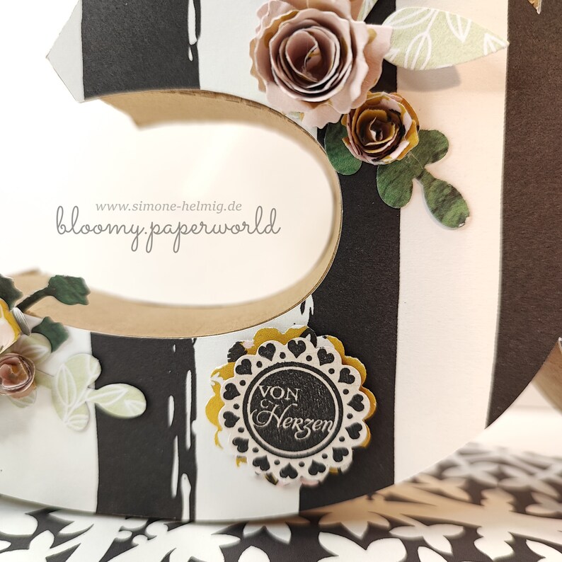 May include: A black and white striped wooden letter 'S' decorated with paper flowers, leaves, and a black and white circular sticker with the text "Von Herzen".