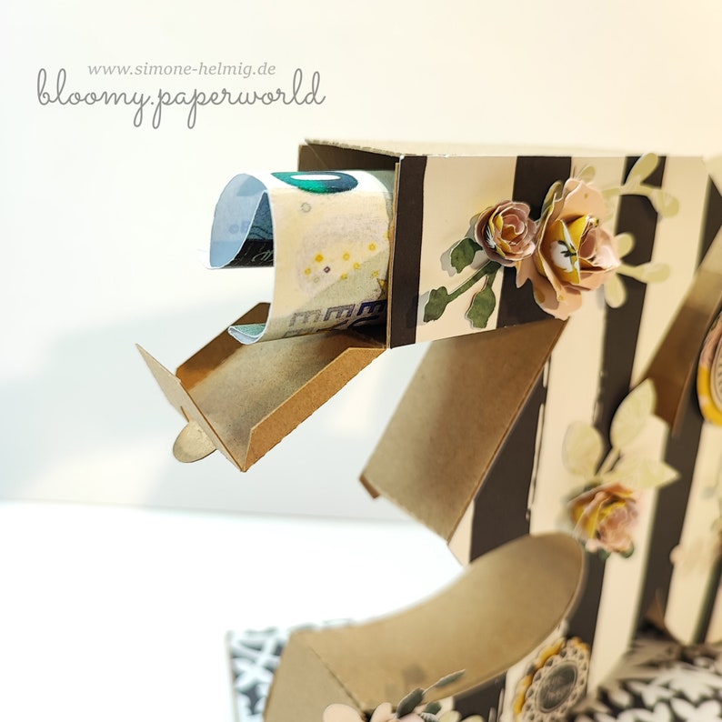 May include: A brown cardboard box with a black and white striped pattern. The box has a small opening with a flap that is open, revealing a rolled up bill of money. The box is decorated with small paper flowers in shades of pink, yellow, and green.