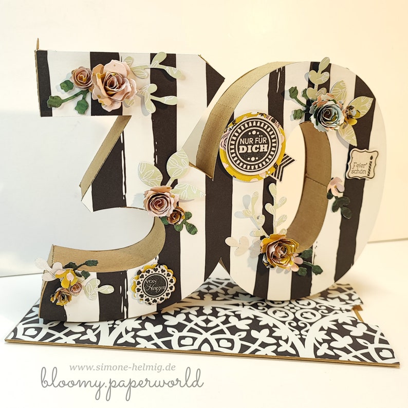May include: A 3D papercraft number 30 with black and white stripes, decorated with flowers, leaves, and paper embellishments. The number is standing on a black and white patterned surface. The text "Nur fur Dich" is on a yellow circle with black dots.