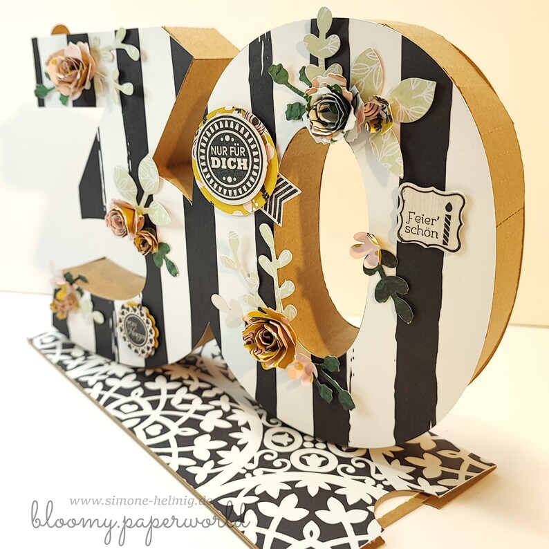 May include: A 3D cardboard number "30" decorated with black and white stripes, paper flowers, and small paper embellishments. The number is standing on a black and white patterned base. The embellishments include the text "Nur f&uuml;r Dich" and "Feier schon".
