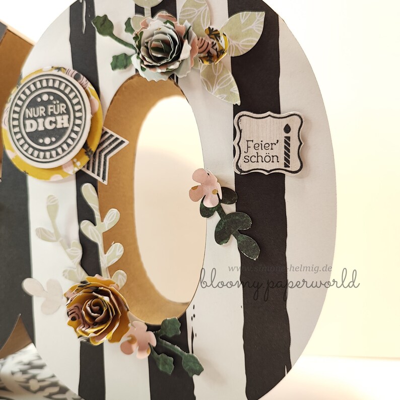 May include: A close-up of a large, brown cardboard letter "O" decorated with black and white stripes, paper flowers, and a small paper tag that says "Feier' sch&ouml;n".
