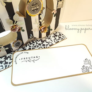 May include: A white card with a brown border and black text that reads "Gl&uuml;ckw&uuml;nsche zum FREUTAG."  The card has a black and white floral design in the bottom right corner. The card is sitting on a black and white patterned surface.