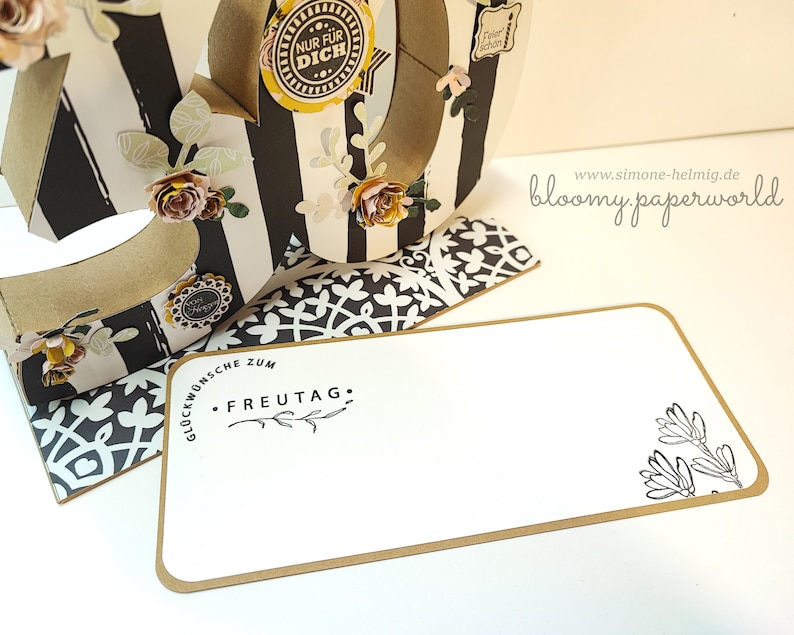 May include: A white card with a brown border and black text that reads "Gl&uuml;ckw&uuml;nsche zum FREUTAG."  The card has a black and white floral design in the bottom right corner. The card is sitting on a black and white patterned surface.