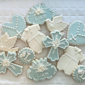 May include: A collection of decorated sugar cookies in light blue and white, featuring shapes of crosses, hearts with rosary beads, doves, baby outfits, and plaques with the name Liam or the letter L, all adorned with intricate white icing details.