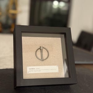 May include: A framed artwork featuring a silver lynchpin on a beige linen background. The black frame surrounds the lynchpin and a small printed definition of the word "Linchpin". The artwork is displayed on a dark surface.