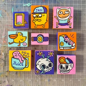 May include: Nine colorful, hand-painted wooden blocks with cartoon-style illustrations. The blocks feature images of characters, a shoe, a camera, a skull, and the word "Juice". Each block is a square shape.