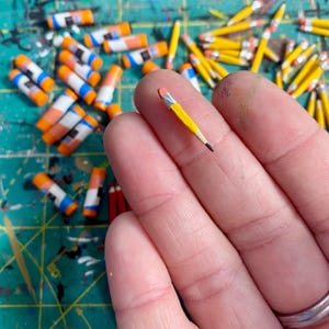 May include: A miniature yellow pencil with a pink eraser and a silver ferrule, featuring a sharpened black lead tip. The tiny pencil is held against a background of numerous other small, similar pencils scattered on a green surface.