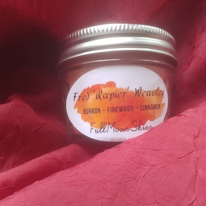 Puede incluir: Una pequeña vela de vidrio en un frasco con una etiqueta blanca que dice "Fred 'Rapier' Weasley" y "Full Moon Skies". La etiqueta también incluye el texto "Bourbon - Firewood - Cinnamon".