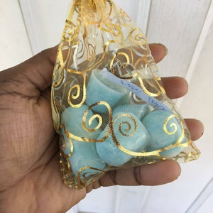 Puede incluir: Una peque&ntilde;a bolsa de malla dorada llena de fundidos de cera azul claro. La bolsa tiene un cord&oacute;n de ajuste dorado y est&aacute; decorada con un patr&oacute;n dorado en espiral.