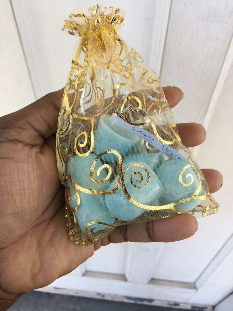 Puede incluir: Una peque&ntilde;a bolsa de malla dorada llena de fundidos de cera azul claro. La bolsa tiene un cord&oacute;n de ajuste dorado y est&aacute; decorada con un patr&oacute;n dorado en espiral.