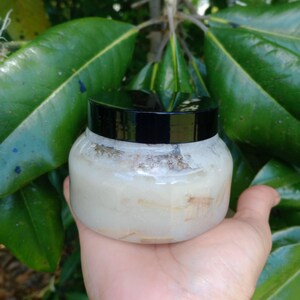 May include: A clear, round jar with a black lid, filled with a creamy, off-white substance. The jar is held in a hand, with green leaves in the background. The substance appears to have small, light brown particles.