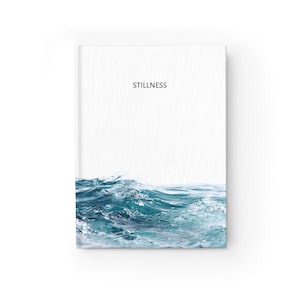 May include: A hardcover journal with a white cover featuring the word STILLNESS printed in black text at the top center. The bottom portion of the cover displays a photographic print of turbulent, deep blue and teal ocean waves with white foam.