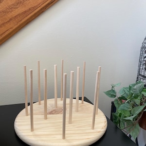 May include: A wooden thread spool holder with a circular base and multiple vertical dowels. The natural wood colour of the base and dowels creates a simple, rustic aesthetic. The holder is designed to organise spools of thread.
