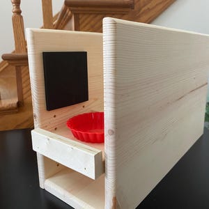 May include: A wooden birdhouse with a natural finish. It features a small, red, scalloped-edge bowl inside and a black square on the front. The birdhouse is constructed from light-colored wood and has a simple, rectangular design.