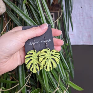 May include: Hand-painted, leaf-shaped earrings in a vibrant yellow and black design. The earrings are held on a black card that reads "HAND-PAINTED". The earrings are hanging in front of a background of green leaves.