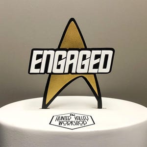 May include: A cake topper with a gold star shape and the word "ENGAGED" in white block letters. The topper is black-edged and sits atop a white cake. The logo for Hunter Valley Workshop is at the base.