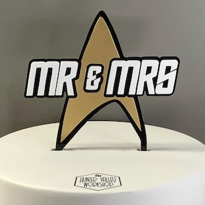 May include: A cake topper featuring the words "MR & MRS" in a bold, white font with a black outline, set against a gold starburst shape. The topper is placed on a white cake. The logo "Hunter Valley Workshop" is at the base.