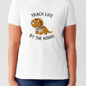 Peut inclure: T-shirt blanc avec un tricératops de dessin animé portant un stéthoscope et le texte "Trach Life By The Horns".