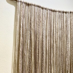 May include: A wall hanging made of yarn in shades of beige, cream, and light pink. The yarn is tied to a piece of driftwood at the top.