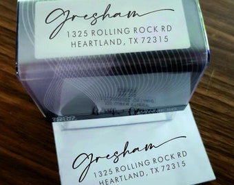 May include: A white self-inking address stamp with a black ink pad. The stamp reads "Gresham 1325 Rolling Rock Rd Heartland, TX 72315".