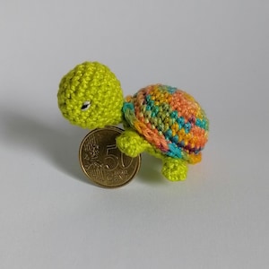 May include: A small, crocheted turtle figurine with a bright lime green head and limbs. The shell features a variegated yarn pattern in shades of orange, blue, and yellow. A 50-cent euro coin is positioned next to the turtle for scale reference.