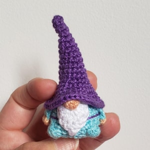 Puede incluir: Pequeña figura de gnomo hecha a mano con ganchillo. El gnomo tiene un gorro puntiagudo morado, una barba blanca y una túnica azul claro. Los brazos y la cara del gnomo son de color melocotón claro. La figura se sostiene en una mano.
